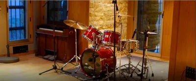 Miked-up red drum kit on a rug beside an upright piano and stone wall